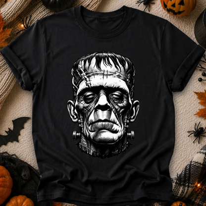 A black short-sleeve crewneck t-shirt featuring a detailed black and white graphic of the character Frankenstein's monster on the front. The shirt is displayed in a flat-lay arrangement with Halloween decorations, including pumpkins, spiderwebs, and a black blanket.