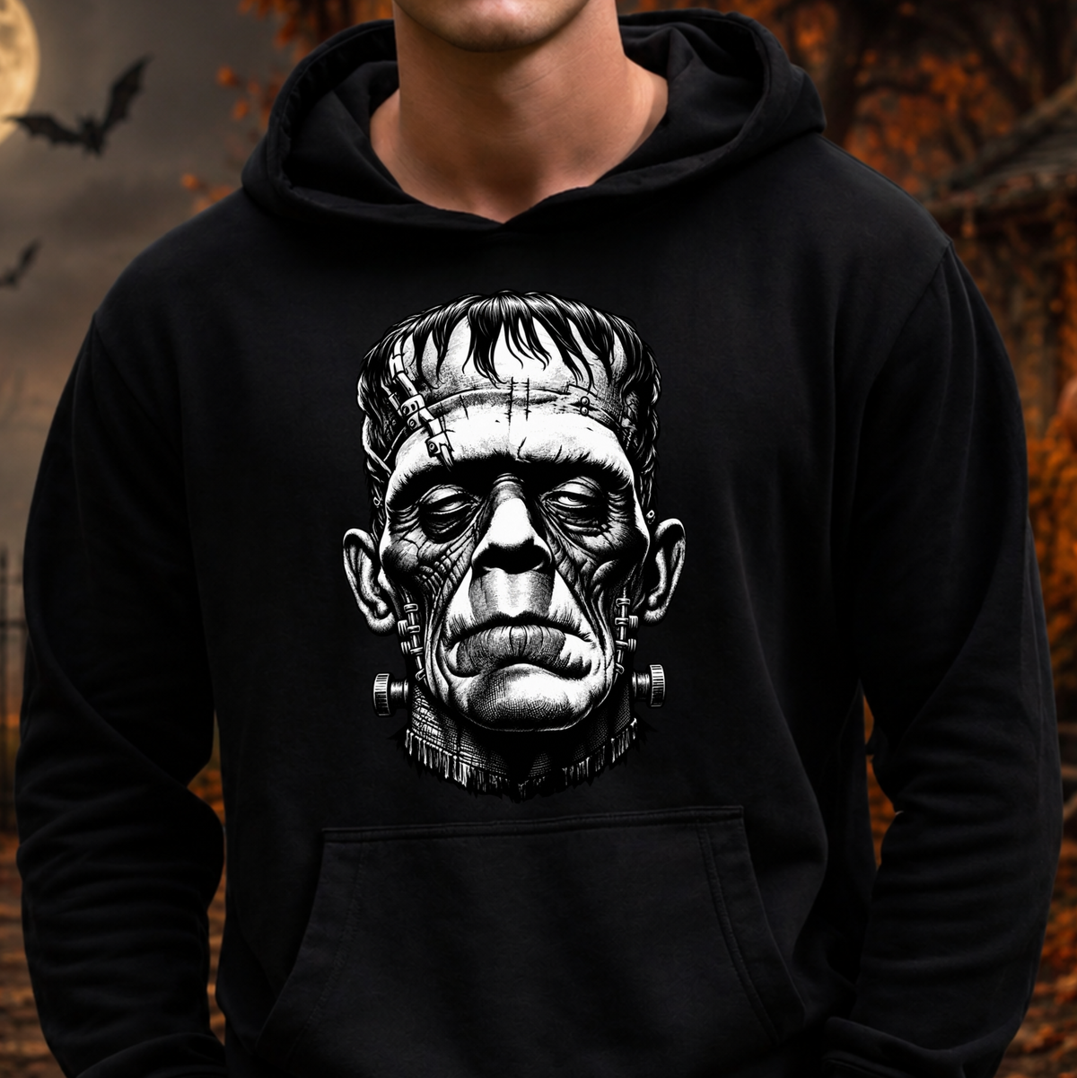 A person wearing a black pullover hoodie with a large black-and-white graphic of Frankenstein's monster face on the chest, set against a dark, spooky outdoor background with bats and autumn leaves.