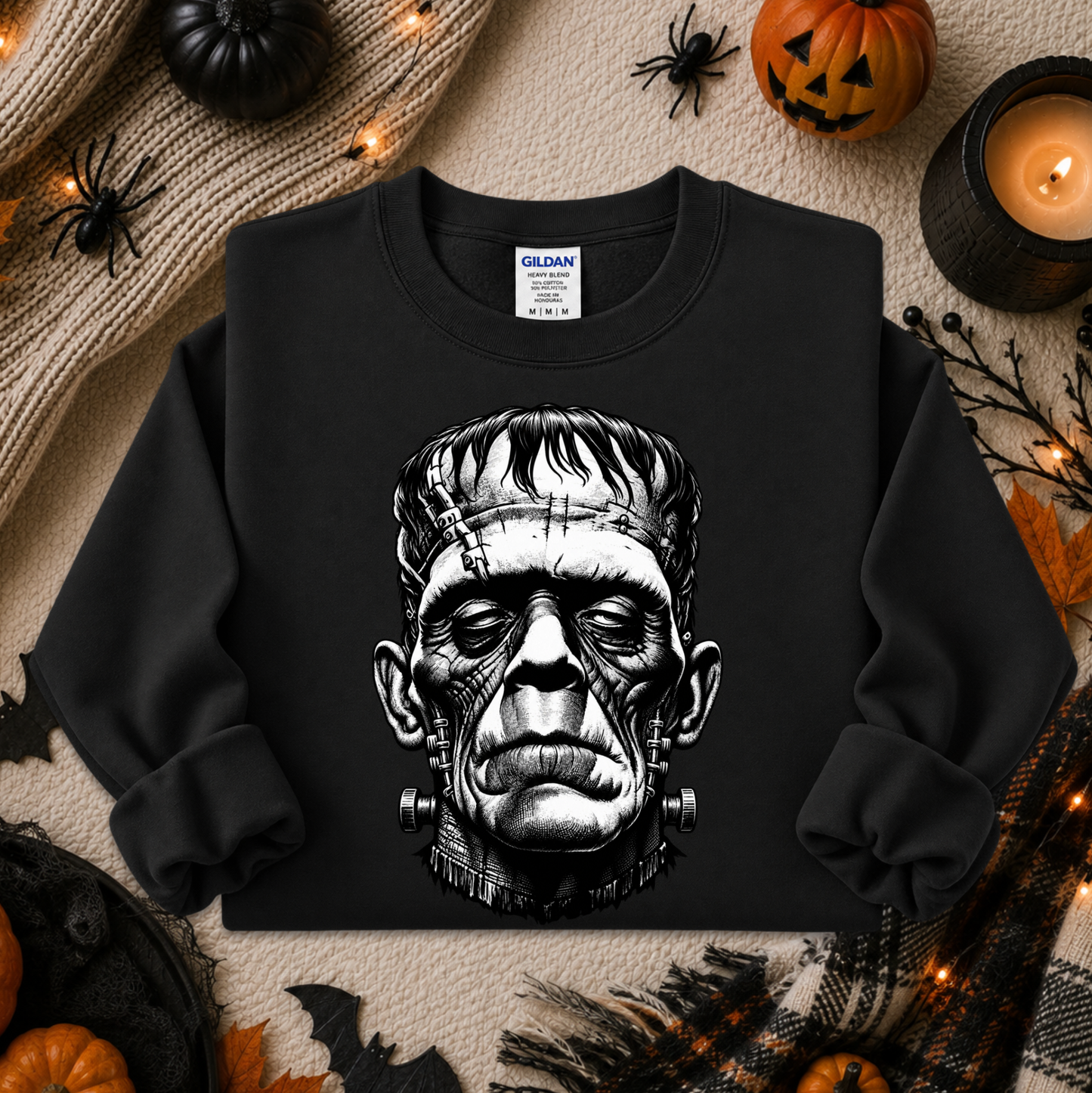 A black crewneck sweatshirt featuring a detailed black and white graphic of Frankenstein's monster head on the front. The sweatshirt is displayed in a flat-lay Halloween setting with pumpkins, a candle, spider decorations, and a cozy knit blanket.