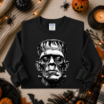 A black crewneck sweatshirt featuring a detailed black and white graphic of Frankenstein's monster head on the front. The sweatshirt is displayed in a flat-lay Halloween setting with pumpkins, a candle, spider decorations, and a cozy knit blanket.