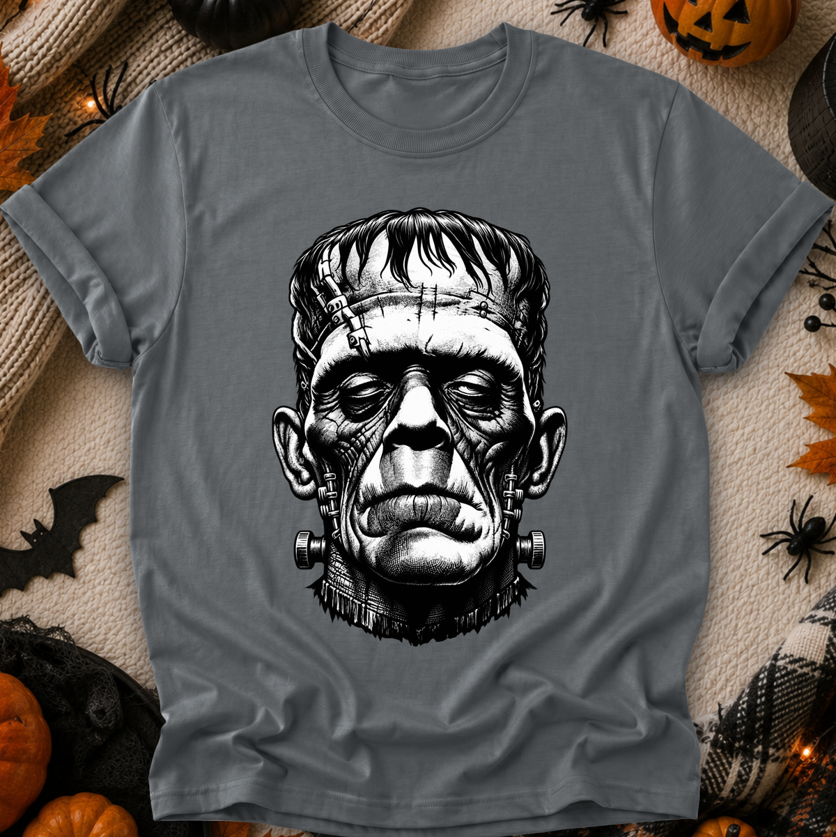A gray short-sleeve crewneck t-shirt featuring a detailed black and white graphic of Frankenstein's monster head on the front. The shirt is displayed in a flat-lay arrangement surrounded by Halloween decorations including pumpkins, a spider, a bat, and autumn leaves on a textured beige blanket.