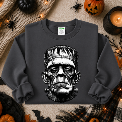 A dark charcoal gray crewneck sweatshirt featuring a detailed black and white graphic of Frankenstein's monster head on the front. The sweatshirt is displayed in a flat-lay arrangement on a cozy beige blanket, surrounded by Halloween decorations including pumpkins, a candle, spiderwebs, and a plaid scarf.
