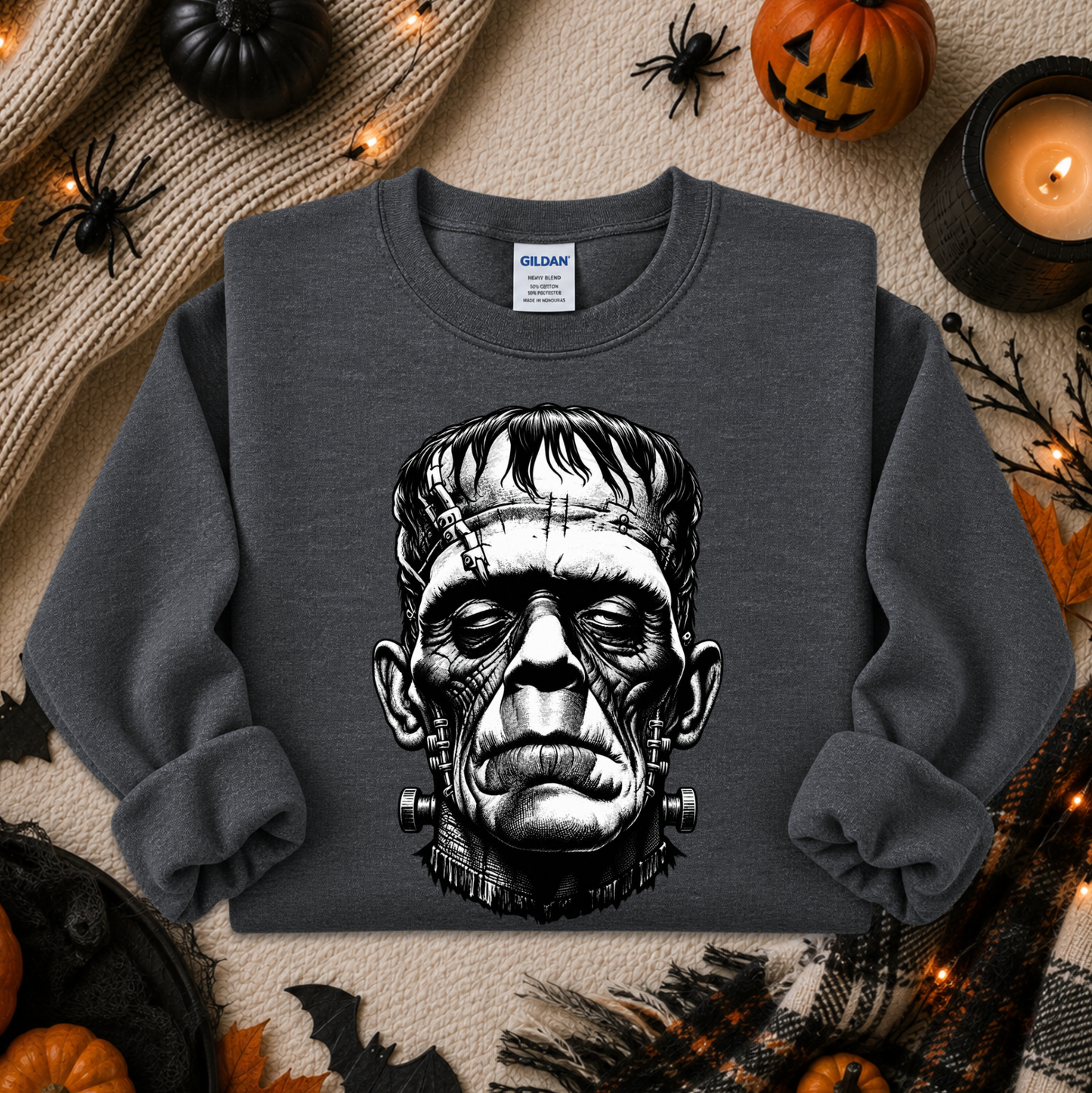 A dark gray crewneck sweatshirt featuring a detailed black and white graphic of Frankenstein's monster head, styled in a flat-lay Halloween setting with pumpkins, a candle, and spider decorations.