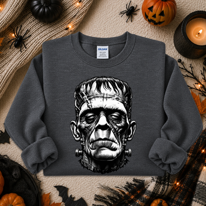 A dark gray crewneck sweatshirt featuring a detailed black and white graphic of Frankenstein's monster head, styled in a flat-lay Halloween setting with pumpkins, a candle, and spider decorations.