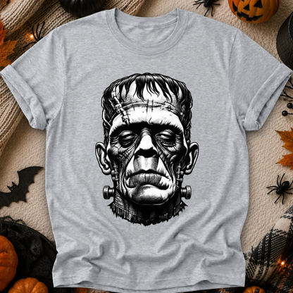 A light gray heather crewneck t-shirt featuring a detailed black and white graphic of Frankenstein's monster, styled in a flat-lay on a cozy beige knit blanket surrounded by Halloween decorations like pumpkins, a spider, and a bat.