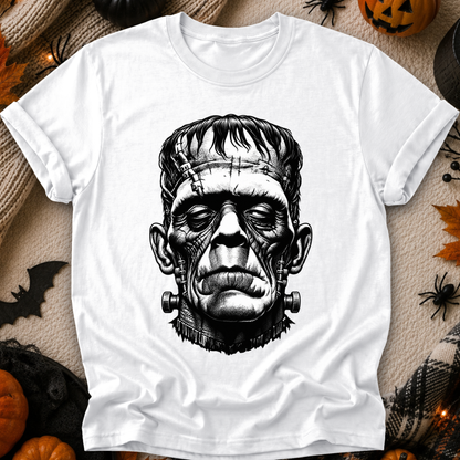 A white short-sleeve crewneck t-shirt featuring a detailed black ink illustration of Frankenstein's monster head on the front, displayed in a flat-lay Halloween setting with pumpkins, a spider, and a black cat.