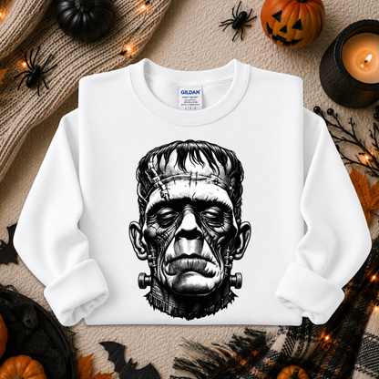 A white long-sleeve crewneck sweatshirt with a black graphic of Frankenstein's monster face, displayed in a flat-lay Halloween setting with pumpkins, a candle, and spider decorations.