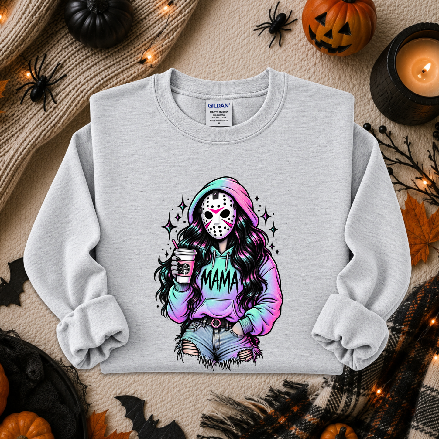 A light gray crewneck sweatshirt with a colorful graphic of a woman in a hockey mask holding a coffee cup and wearing a hoodie that says 'MAMA'. The sweatshirt is displayed in a flat-lay Halloween setting with pumpkins, a candle, and spider decorations.
