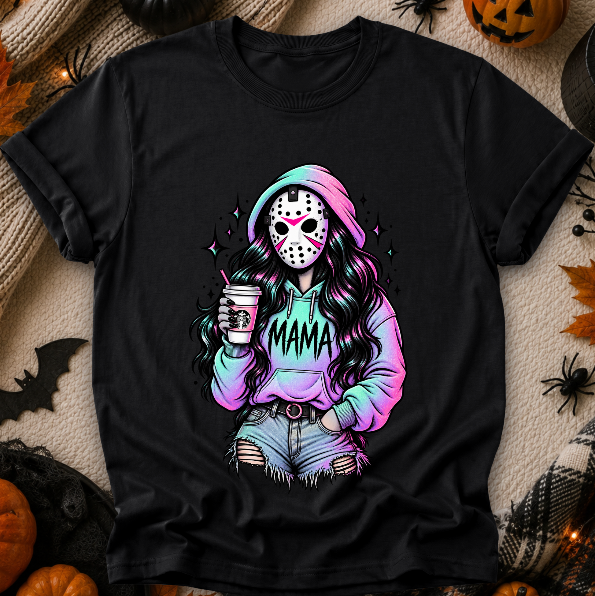 A black short-sleeve graphic T-shirt laid flat on a cozy beige fabric background with Halloween props like pumpkins, spider decorations, and a bat. The shirt features a colorful printed illustration of a woman in a hockey mask holding a drink, with the word "MAMA" across the hoodie.