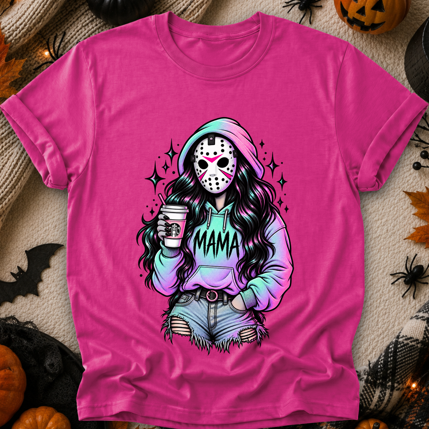 A bright pink graphic t-shirt laid flat on a cozy autumn-themed background with pumpkins, spider decorations, and a knit blanket. The graphic features a stylized horror character resembling Jason Voorhees with long dark hair, a hockey mask, and a hoodie that says 'MAMA'.