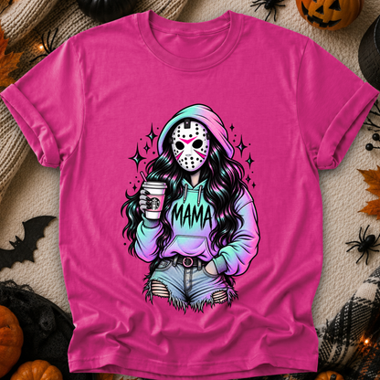 A bright pink graphic t-shirt laid flat on a cozy autumn-themed background with pumpkins, spider decorations, and a knit blanket. The graphic features a stylized horror character resembling Jason Voorhees with long dark hair, a hockey mask, and a hoodie that says 'MAMA'.