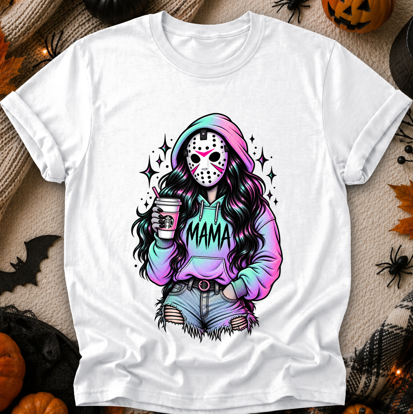 A white short-sleeve graphic T-shirt laid flat on a cozy autumn-themed background with pumpkins, spider decorations, and faux fur. The shirt features a colorful illustration of a woman in a hockey mask and pastel rainbow hoodie with the word 'MAMA' across the front.