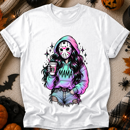 A white short-sleeve graphic T-shirt laid flat on a cozy autumn-themed background with pumpkins, spider decorations, and faux fur. The shirt features a colorful illustration of a woman in a hockey mask and pastel rainbow hoodie with the word 'MAMA' across the front.