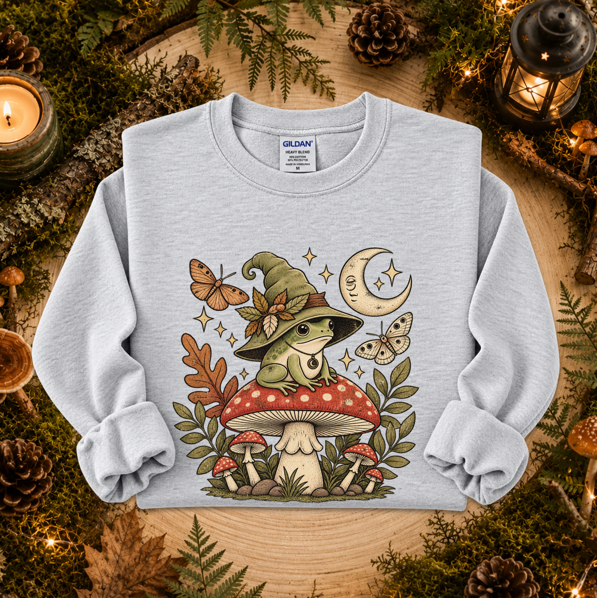 A light gray heather crewneck sweatshirt featuring a whimsical graphic of a green frog wearing a witch hat on a red mushroom, surrounded by butterflies, a crescent moon, and autumn leaves. The sweatshirt is displayed flat on a rustic wooden surface decorated with pinecones, moss, and candles.
