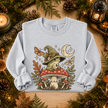 A light gray heather crewneck sweatshirt featuring a whimsical graphic of a green frog wearing a witch hat on a red mushroom, surrounded by butterflies, a crescent moon, and autumn leaves. The sweatshirt is displayed flat on a rustic wooden surface decorated with pinecones, moss, and candles.