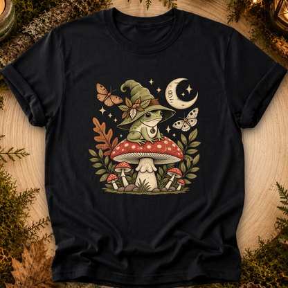 A black short-sleeve crewneck t-shirt featuring a whimsical graphic of a green frog wearing a witch hat, sitting on a red and white mushroom with smaller mushrooms, butterflies, stars, and a crescent moon around it. The shirt is displayed flat on a rustic wood background decorated with pine needles and dried leaves.