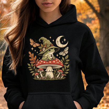 A woman wearing a black hoodie with a detailed graphic of a green frog in a witch hat sitting on a red mushroom, surrounded by butterflies, stars, and autumn leaves, set against a blurred autumn forest background.