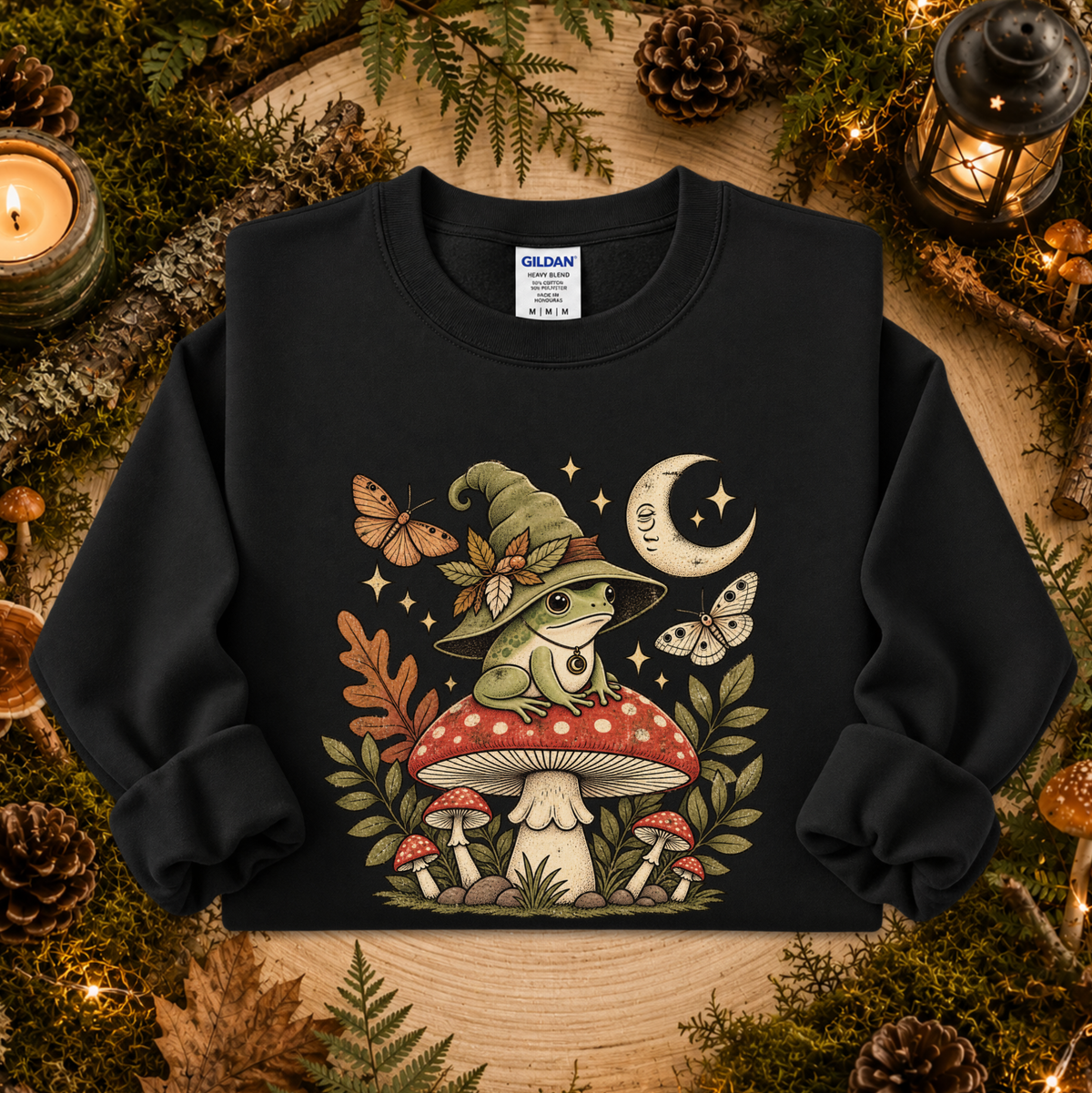 A black crewneck sweatshirt featuring a detailed graphic of a green frog wearing a witch hat sitting on a red and white mushroom, surrounded by butterflies, stars, and a crescent moon. The sweatshirt is displayed in a cozy, autumn-themed flat lay with pinecones, moss, and candles.