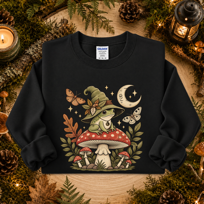 A black crewneck sweatshirt featuring a detailed graphic of a green frog wearing a witch hat sitting on a red and white mushroom, surrounded by butterflies, stars, and a crescent moon. The sweatshirt is displayed in a cozy, autumn-themed flat lay with pinecones, moss, and candles.