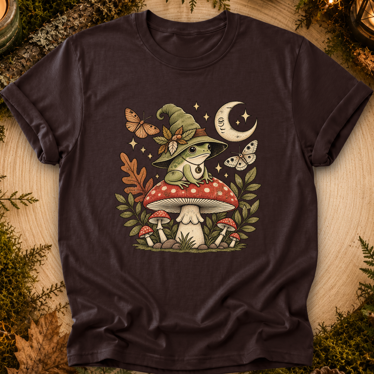 A dark brown short-sleeve t-shirt with a whimsical graphic of a green frog wearing a witch hat, sitting on a red and white mushroom. The design is surrounded by autumn elements including a crescent moon, butterflies, stars, and smaller mushrooms, all set against a background of wood and green foliage.