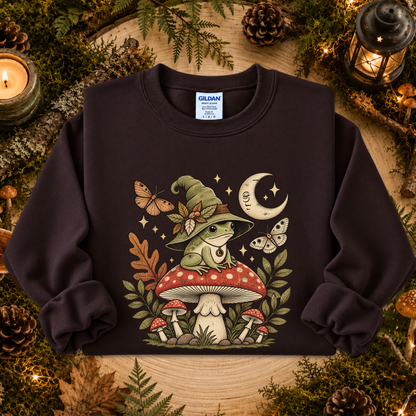 A dark brown crewneck sweatshirt by Gildan, featuring a whimsical graphic of a green frog wizard sitting on a red and white mushroom, surrounded by butterflies, stars, and a crescent moon. The sweatshirt is displayed in a cozy, rustic flat-lay setting with pinecones, moss, and a lit candle.