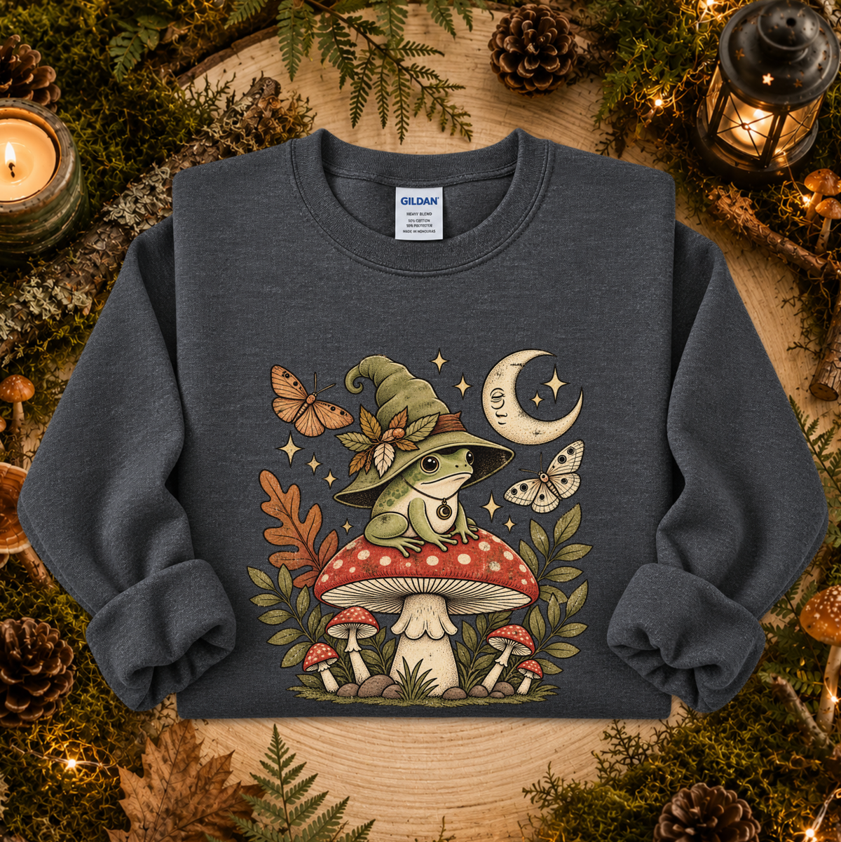 A dark heather gray crewneck sweatshirt featuring a whimsical graphic of a green frog in a witch hat sitting on a red mushroom, surrounded by butterflies, stars, and smaller mushrooms. The sweatshirt is displayed flat on a rustic wooden background decorated with pinecones, moss, and candles.