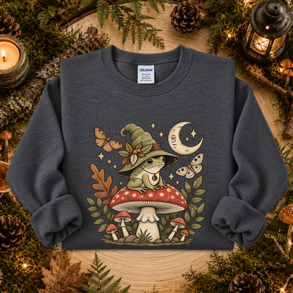 A dark heather gray crewneck sweatshirt featuring a whimsical graphic of a green frog in a witch hat sitting on a red mushroom, surrounded by butterflies, stars, and smaller mushrooms. The sweatshirt is displayed flat on a rustic wooden background decorated with pinecones, moss, and candles.