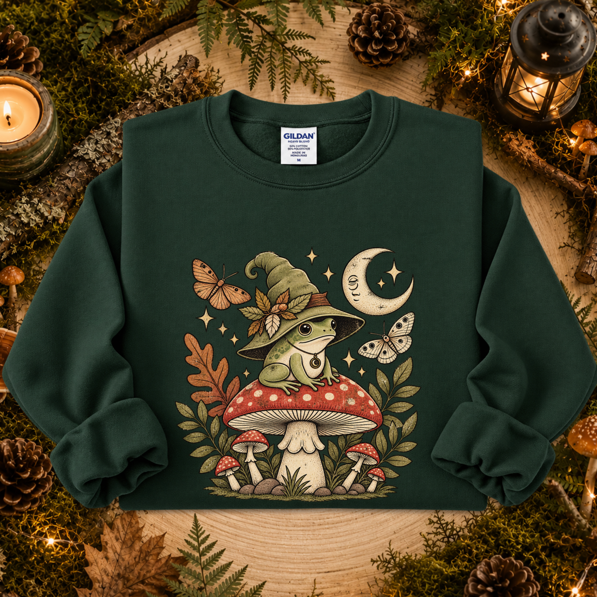 A dark green crewneck sweatshirt featuring a whimsical graphic of a green frog in a witch hat sitting on a red-and-white mushroom, surrounded by butterflies, stars, and a crescent moon. The sweatshirt is displayed in a cozy flat-lay setting with pinecones, moss, and candles.