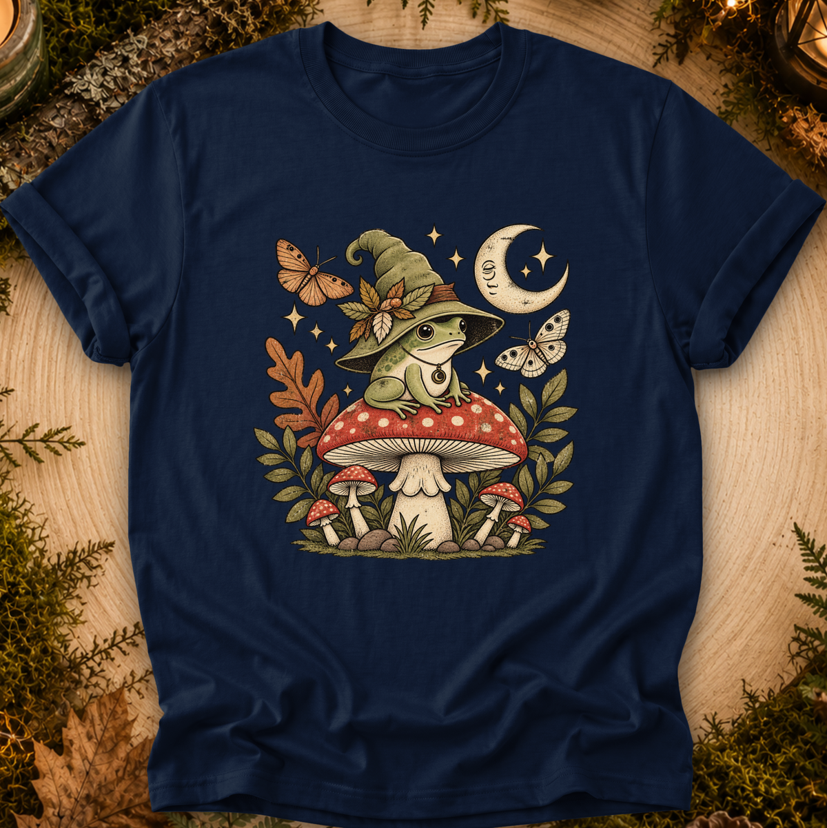 A navy blue short-sleeve t-shirt with a detailed graphic of a frog in a witch hat sitting on a red mushroom, surrounded by butterflies, stars, a crescent moon, and other mushrooms, displayed on a rustic wood background with greenery.