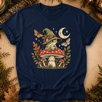A navy blue short-sleeve t-shirt with a detailed graphic of a frog in a witch hat sitting on a red mushroom, surrounded by butterflies, stars, a crescent moon, and other mushrooms, displayed on a rustic wood background with greenery.