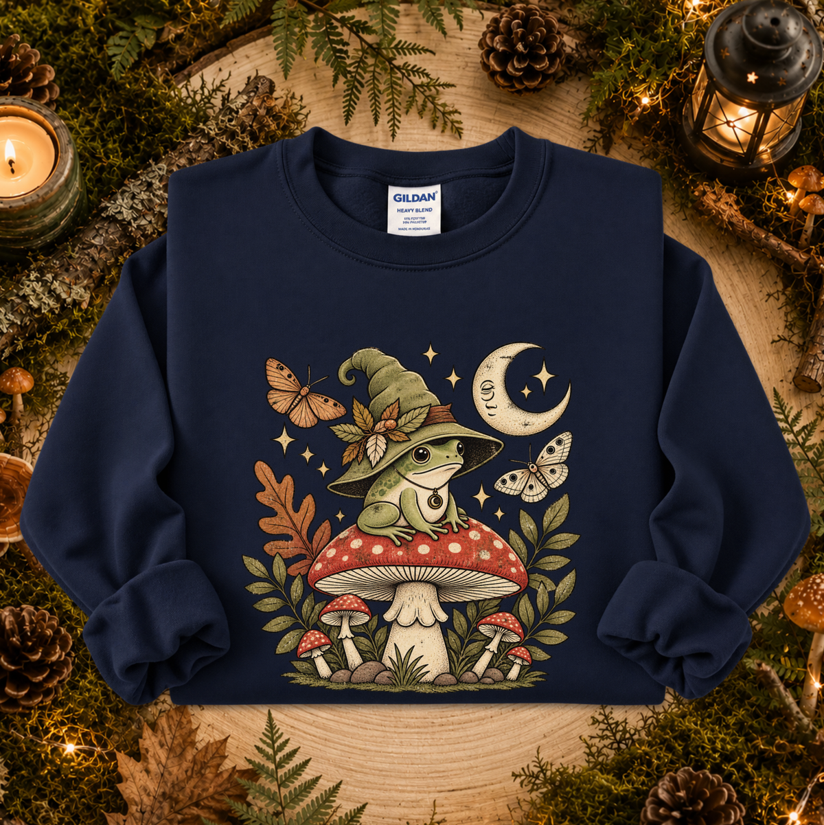 A navy blue Gildan sweatshirt featuring a whimsical graphic of a green frog wearing a witch hat on a red mushroom, surrounded by butterflies, stars, and a crescent moon, displayed in a cozy autumn-themed flat lay with candles, pinecones, and moss.