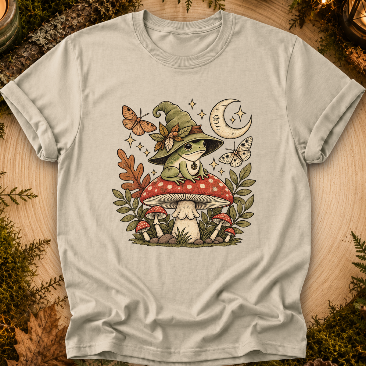 A light beige crewneck t-shirt featuring a whimsical graphic of a green frog wearing a witch hat sitting on a red and white mushroom, surrounded by butterflies, leaves, and a crescent moon, displayed on a rustic wood background with moss.