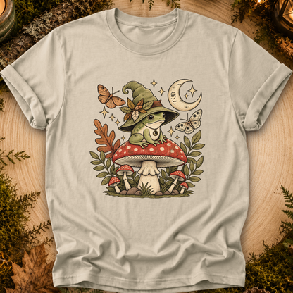 A light beige crewneck t-shirt featuring a whimsical graphic of a green frog wearing a witch hat sitting on a red and white mushroom, surrounded by butterflies, leaves, and a crescent moon, displayed on a rustic wood background with moss.