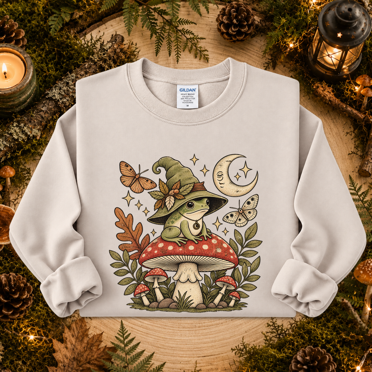 A light beige crewneck sweatshirt featuring a whimsical graphic of a green frog in a witch hat sitting on a red mushroom, surrounded by butterflies, stars, and other mushrooms. The sweatshirt is displayed flat on a rustic wooden surface decorated with pinecones, moss, and candles.
