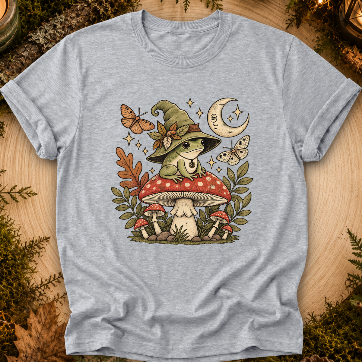 A light heather gray short-sleeve t-shirt with a detailed graphic of a green frog in a witch hat sitting on a red and white mushroom, surrounded by butterflies, leaves, and a crescent moon, displayed flat on a rustic wood background with moss accents.