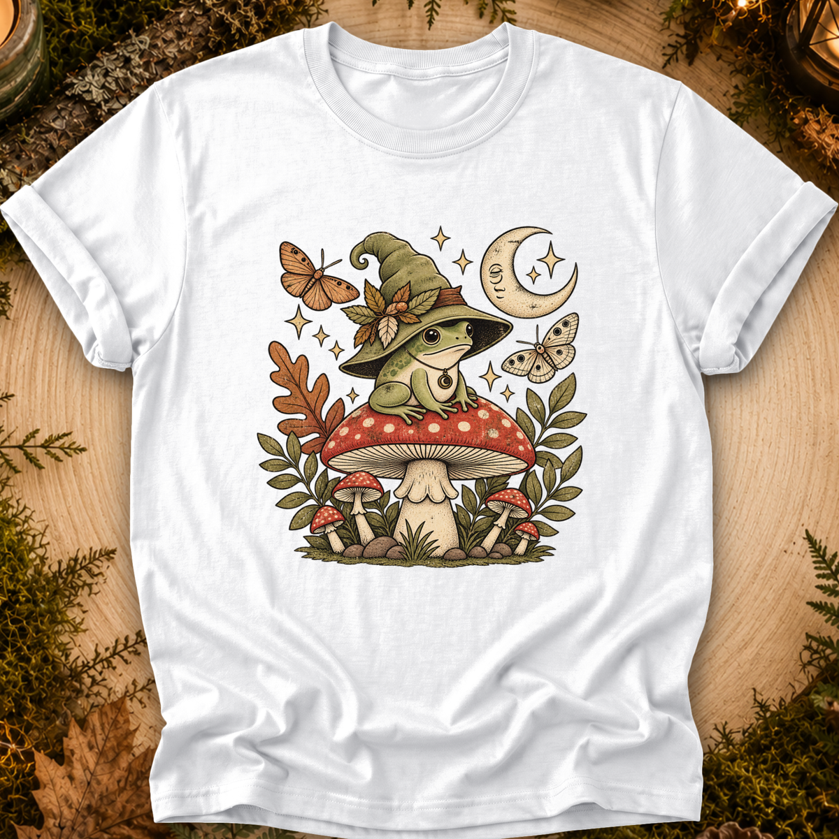 A white short-sleeve crewneck t-shirt laid flat on a rustic wood background with moss and leaves. The shirt features a detailed graphic of a green frog wearing a witch hat, sitting on a red and white mushroom, surrounded by butterflies, stars, and a crescent moon.