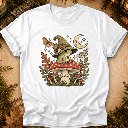 A white short-sleeve crewneck t-shirt laid flat on a rustic wood background with moss and leaves. The shirt features a detailed graphic of a green frog wearing a witch hat, sitting on a red and white mushroom, surrounded by butterflies, stars, and a crescent moon.