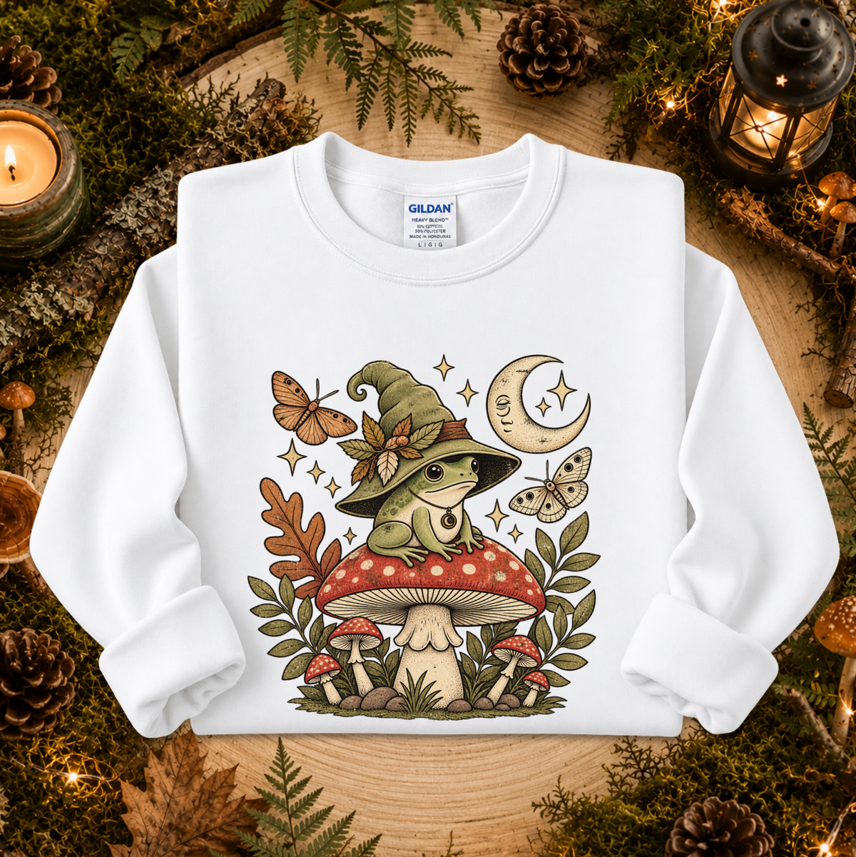 A white long-sleeve sweatshirt laid flat on a rustic wood surface surrounded by moss, pinecones, candles, and lanterns. The front has a large illustrated graphic of a frog wearing a witch hat sitting on a red-and-white mushroom, with butterflies, stars, leaves, and a crescent moon.