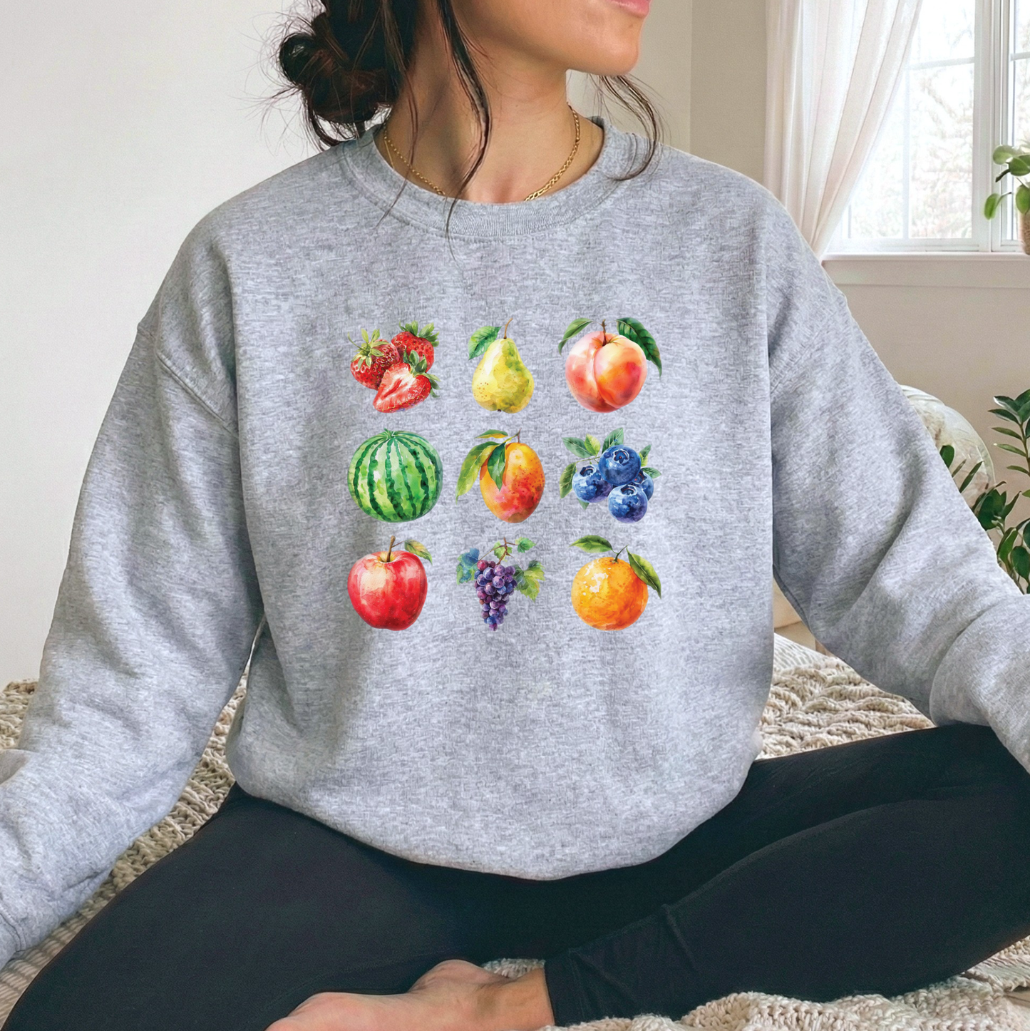 A woman wearing a heather gray crewneck sweatshirt with a colorful watercolor fruit print on the front, sitting on a textured blanket in a bright room.