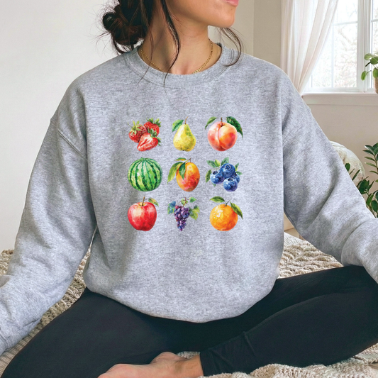 A woman wearing a heather gray crewneck sweatshirt with a colorful watercolor fruit print on the front, sitting on a textured blanket in a bright room.