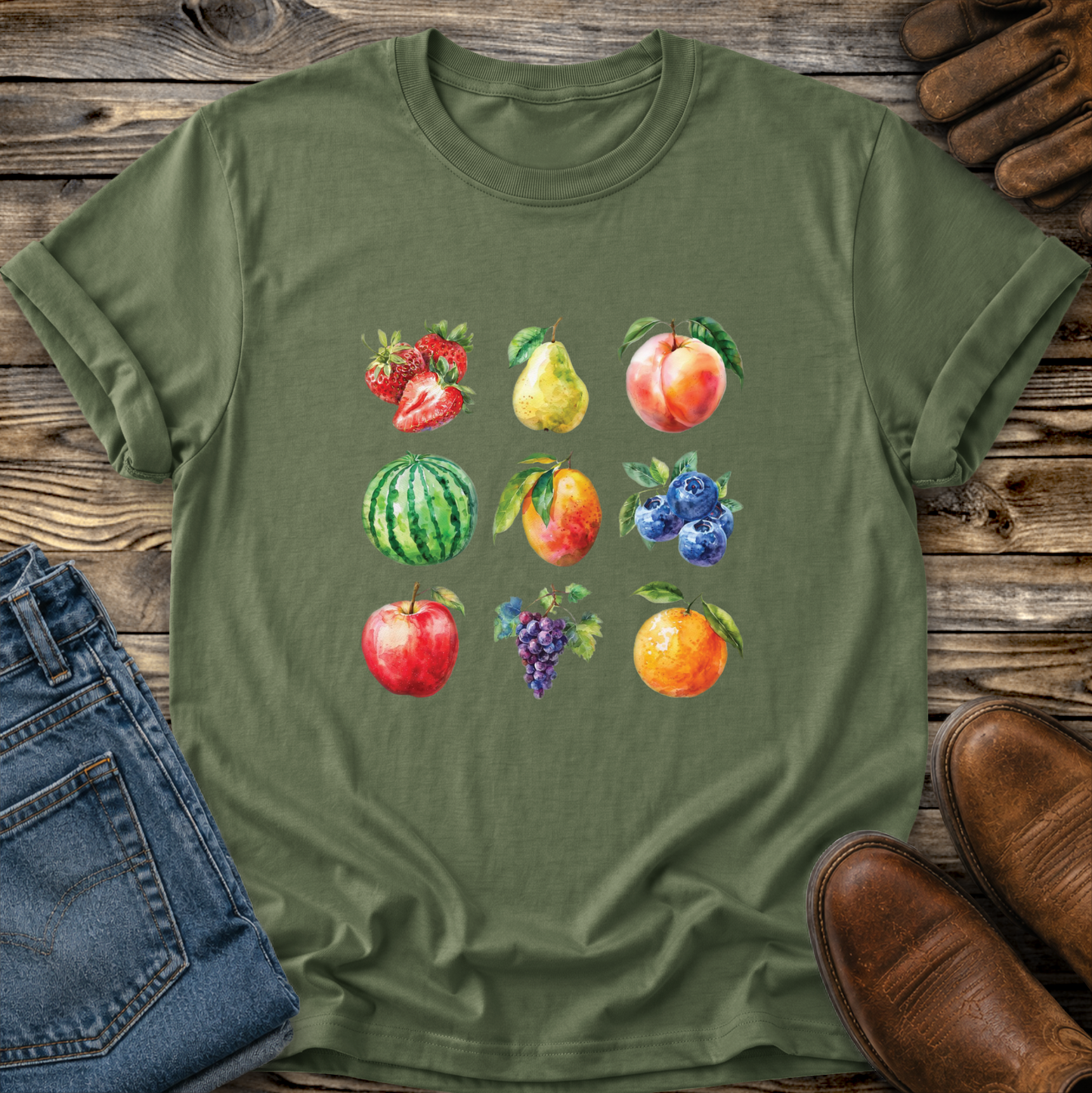 An olive green short-sleeve t-shirt with a colorful watercolor print of various fruits (strawberries, pear, peach, watermelon, blueberries, apple, grapes, and orange) on the front, styled in a flat-lay arrangement on a rustic wooden background with blue jeans and brown leather shoes.