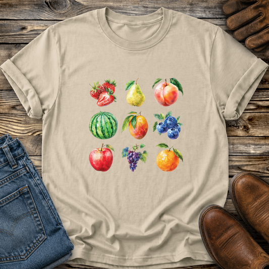 A beige short-sleeve crewneck t-shirt featuring a colorful watercolor print of various fruits (strawberries, pear, peach, watermelon, blueberries, apple, grapes, orange) arranged in a grid. The t-shirt is styled in a flat-lay on a rustic wooden surface, surrounded by blue jeans, brown leather shoes, and leather sandals.
