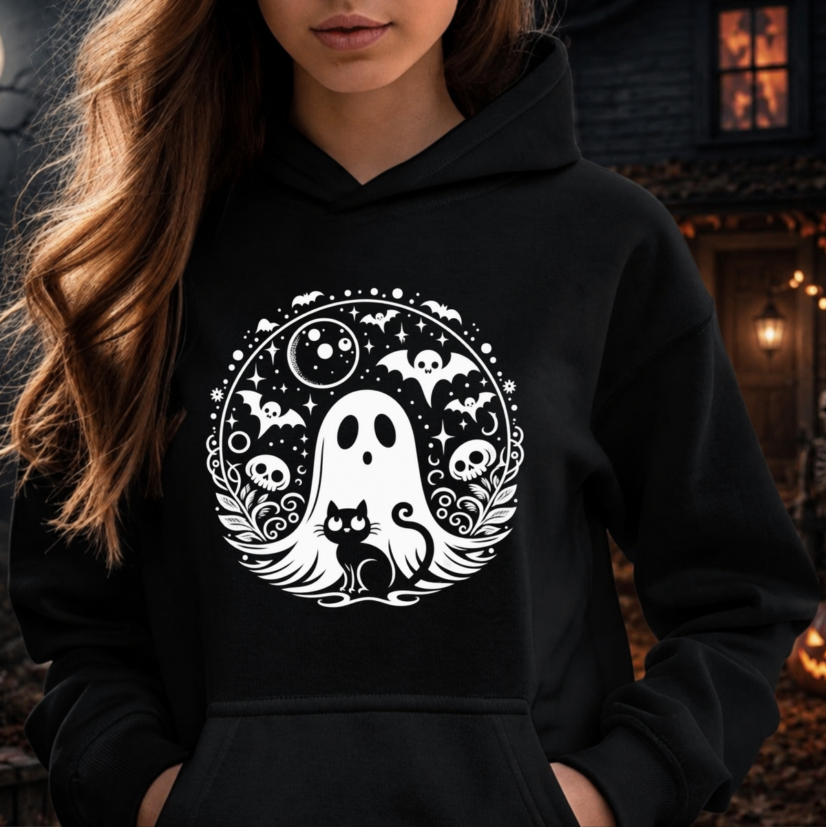 A woman wearing a black hoodie with a white circular Halloween graphic featuring a ghost, cat, skulls, bats, and stars, photographed outdoors in front of a dimly lit house.