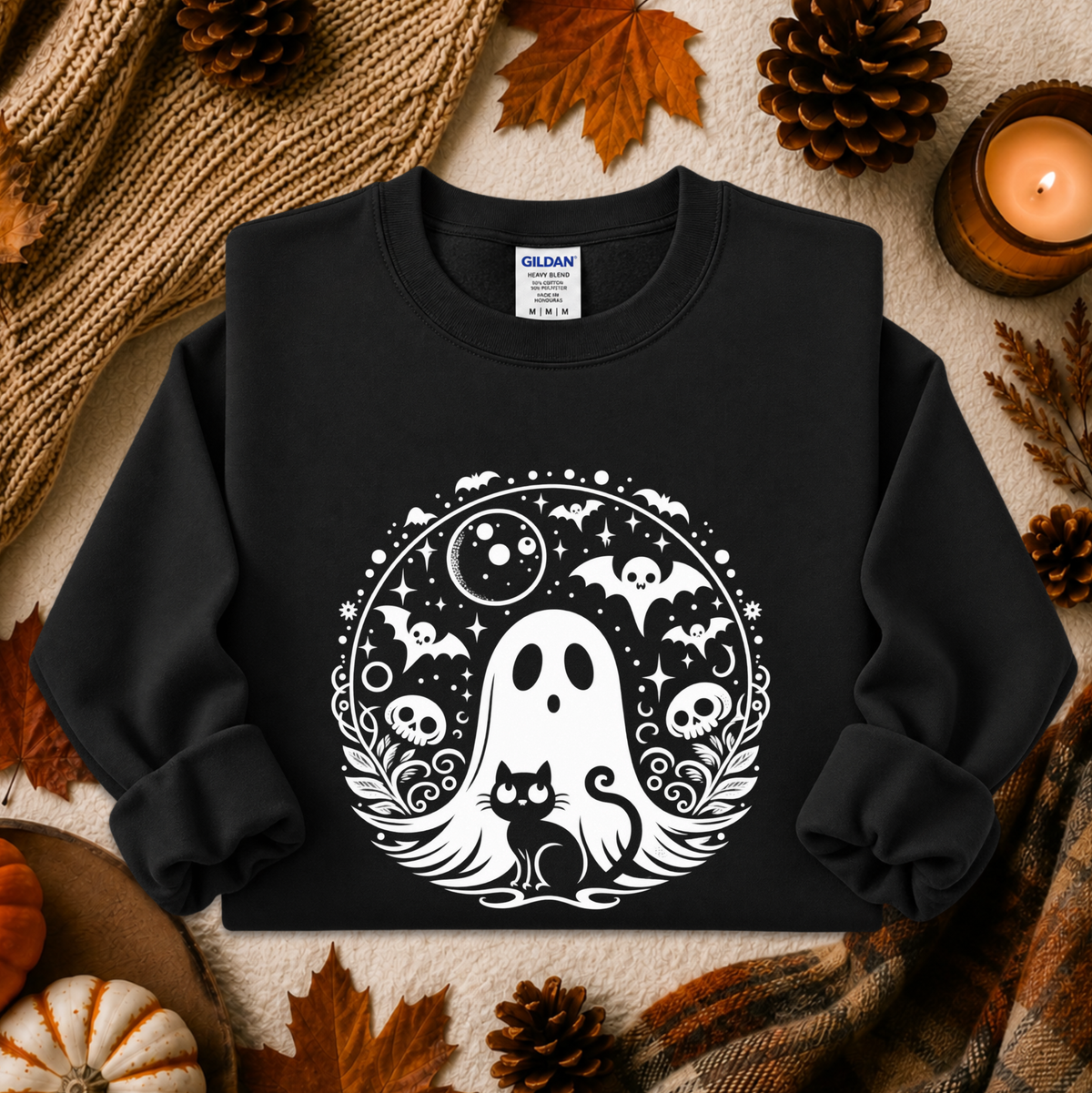 A black crewneck sweatshirt with a white circular graphic of a ghost, cat, bats, skulls, and moon, styled in an autumn flat-lay with pinecones, leaves, a candle, and a knit blanket.