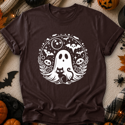 A dark brown short-sleeve t-shirt with a white Halloween graphic print featuring a ghost, cat, bats, skulls, and moons, laid flat on a textured beige blanket surrounded by seasonal props like pumpkins, spider decorations, and autumn leaves.