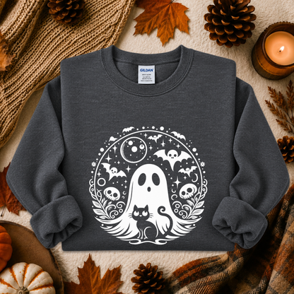 A dark charcoal gray crewneck sweatshirt with a white and black Halloween graphic of a ghost, cat, bats, and skulls, styled in a flat lay with autumn leaves, pinecones, a candle, and a knit blanket.