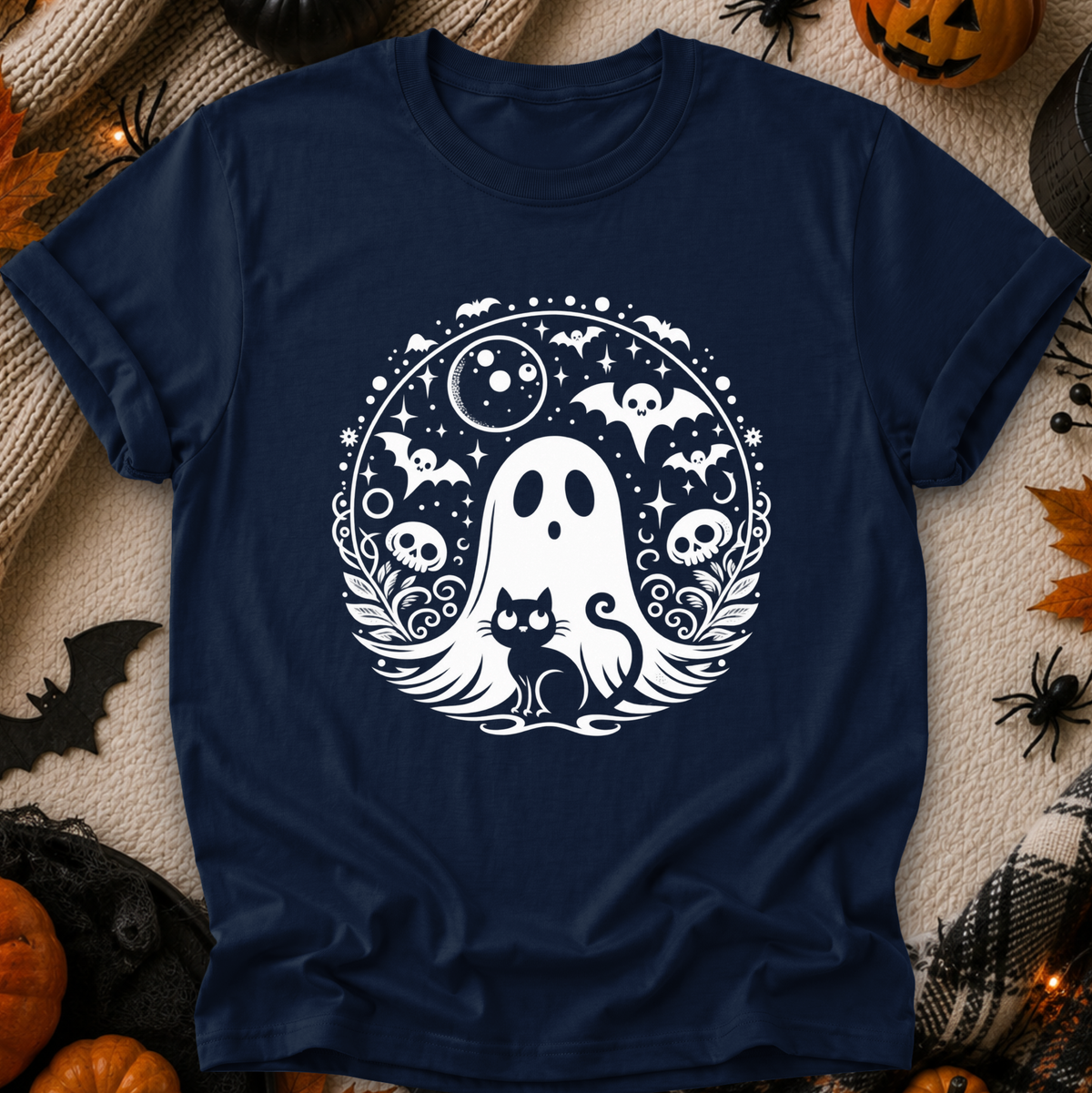 A navy blue short-sleeve t-shirt with a white Halloween graphic of a ghost, cat, bats, skulls, and stars, laid flat on a textured beige blanket with seasonal props like pumpkins and spider decorations.
