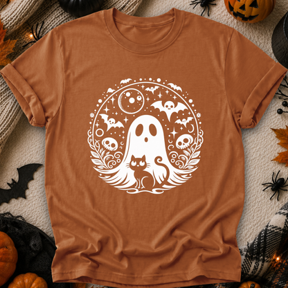 A rust-orange crewneck t-shirt with a white Halloween graphic of a ghost, cat, bats, and skulls, displayed flat on a cozy autumn-themed background with pumpkins, spider decorations, and a knit blanket.