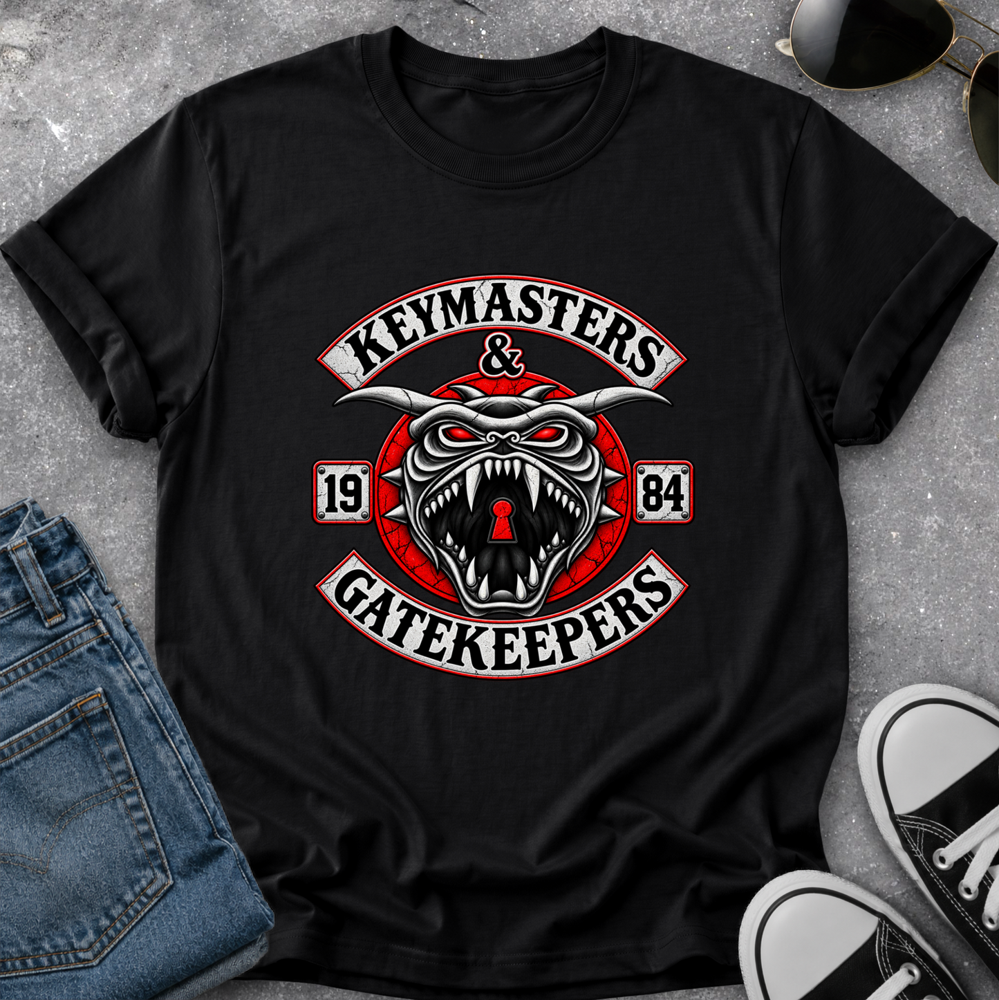A black short-sleeve graphic T-shirt laid flat on a gray textured surface, styled with blue jeans, sunglasses, and black-and-white sneakers. The shirt features a large central graphic with the words 'Keymasters & Gatekeepers' and a snarling horned creature with red eyes and a red circular emblem.