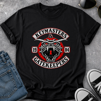 A black short-sleeve graphic T-shirt laid flat on a gray textured surface, styled with blue jeans, sunglasses, and black-and-white sneakers. The shirt features a large central graphic with the words 'Keymasters & Gatekeepers' and a snarling horned creature with red eyes and a red circular emblem.